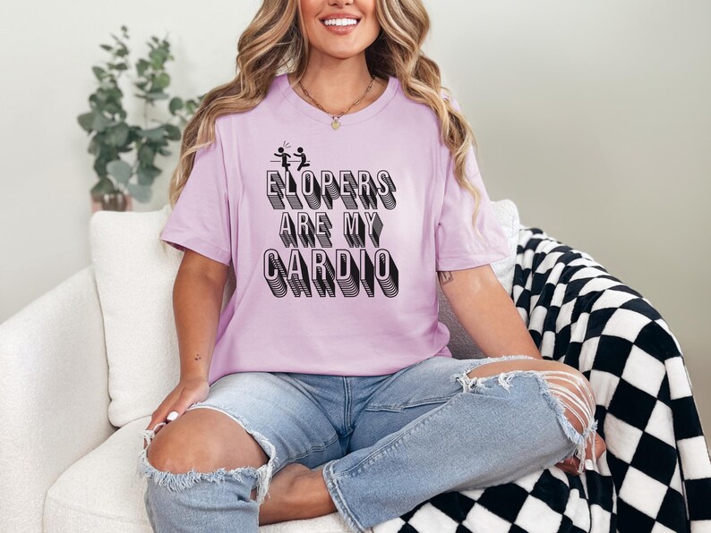 May include: A woman wearing a light purple t-shirt with the text "ELOPERS ARE MY CARDIO" in black. The text is in a 3D style with a black outline. She is sitting on a white couch with a black and white checkered throw blanket.