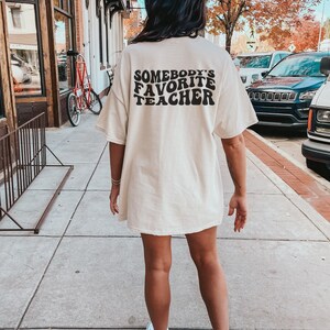 May include: A woman wearing a light beige t-shirt with the text "Somebody's Favorite Teacher" in black. The text is in a retro font style.