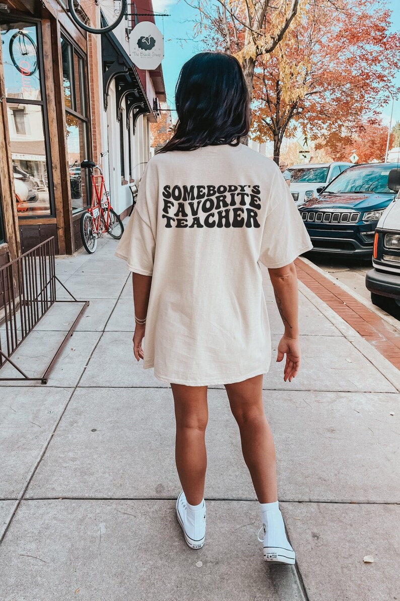 May include: A woman wearing a light beige t-shirt with the text "Somebody's Favorite Teacher" in black. The text is in a retro font style.