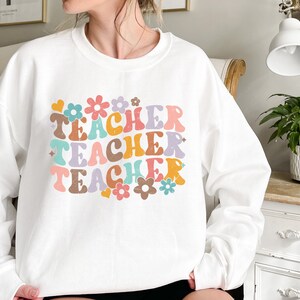 May include: White sweatshirt with the words "Teacher Teacher Teacher" in colorful retro font with flowers and hearts around the text.