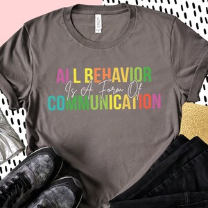 Op de afbeelding: Een grijs T-shirt met de tekst "All Behavior Is A Form Of Communication" gedrukt in regenboogkleuren.