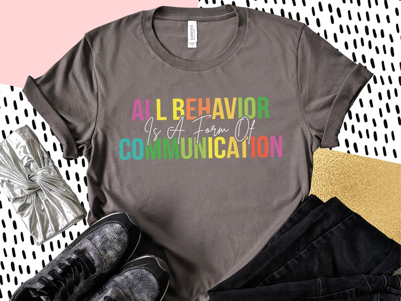 Op de afbeelding: Een grijs T-shirt met de tekst "All Behavior Is A Form Of Communication" gedrukt in regenboogkleuren.