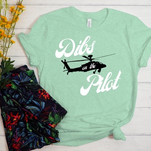 May include: A light green t-shirt with a white graphic of a helicopter and the text "Dibs on the Pilot" in a retro font.