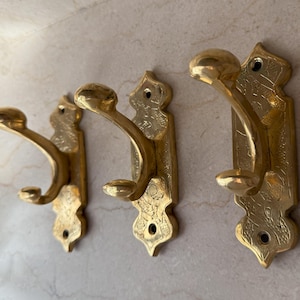 May include: Three antique brass wall hooks. Each hook has a curved design with a rounded knob at the end. The hooks are mounted on decorative backplates with etched patterns and mounting holes.
