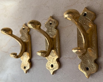 May include: Three antique brass wall hooks. Each hook has a curved design with a rounded knob at the end. The hooks are mounted on decorative backplates with etched patterns and mounting holes.