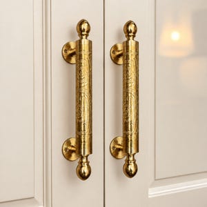 May include: Two vertical brass door handles with a cylindrical shape and a polished gold finish. Each handle features an intricate engraved floral pattern along the center and rounded finials at the top and bottom, mounted with circular plates.