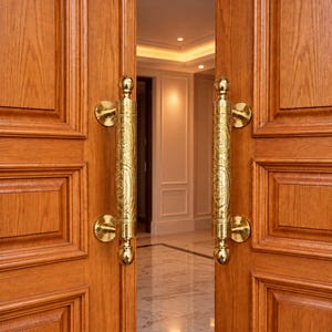 May include: A pair of double wooden doors featuring a natural brown wood grain finish and recessed rectangular panels. Each door is fitted with a vertical, cylindrical brass handle that displays an intricate, embossed floral scroll pattern.