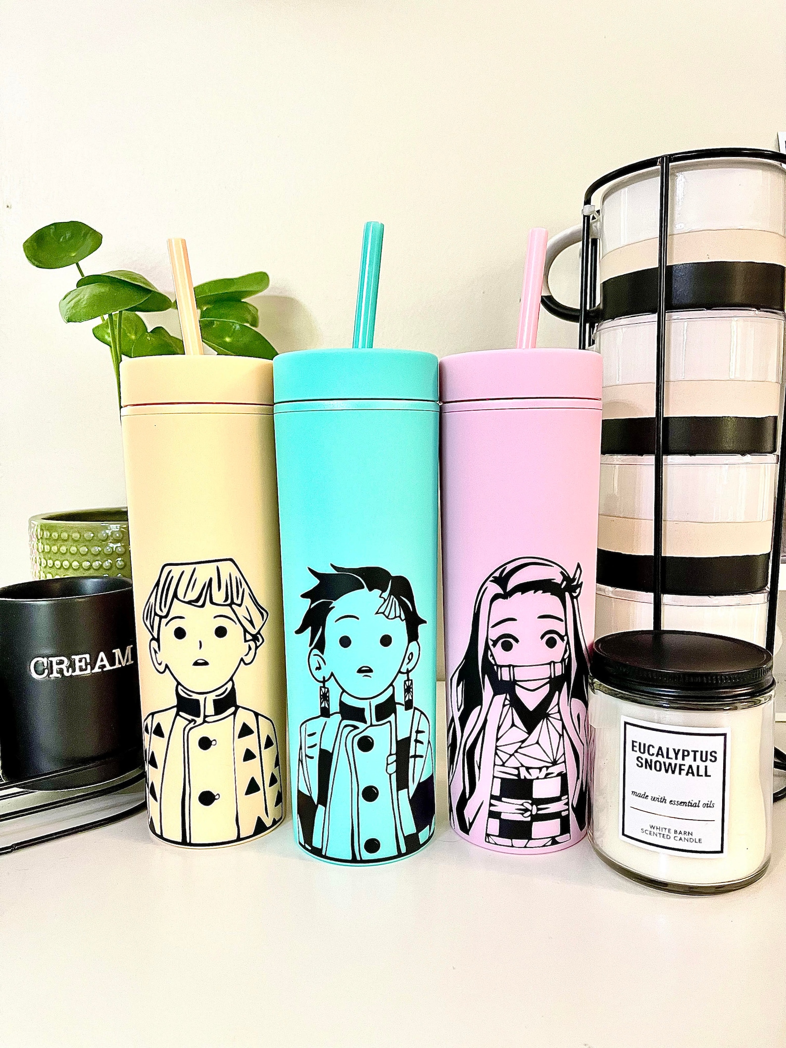 three tumblers. From left to right, they are yellow, turquoise, and pink with Zenitsu, Tanjiro, and Nezuko drawn on each one, respectively