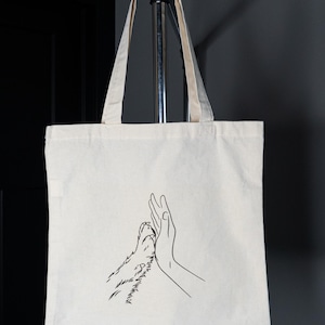 May include: Cream-colored tote bag with a black line drawing of a human hand and a dog paw touching. The bag has long, beige handles and is hanging on a metal rod. The background is a dark gray wall and a black door.