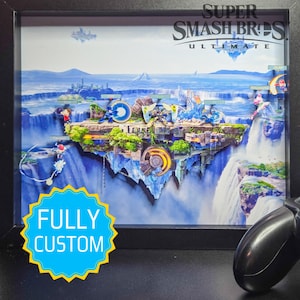 Peut inclure: Un diorama personnalisé et encadré de Super Smash Bros. Ultimate. L'œuvre représente une scène détaillée et superposée d'un niveau de jeu vidéo avec des cascades, des personnages et le texte "FULLY CUSTOM". Le cadre est noir, et une manette de jeu est visible.