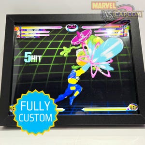 May include: Framed artwork featuring a vibrant scene from the Marvel vs. Capcom video game. The image showcases pixelated characters in action, with the text "5 HIT" and "FULLY CUSTOM" visible. The frame is black.