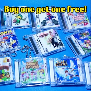 May include: A collection of miniature video game case keychains featuring various Nintendo titles including Mario, Sonic, Animal Crossing, and Kid Icarus. Each clear plastic case displays cover art and includes a metal ball chain. Text reads: Buy one get one free!