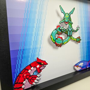 Puede incluir: Una impresi&oacute;n de arte de p&iacute;xeles enmarcada con tres personajes de Pok&eacute;mon: un Pok&eacute;mon de tipo agua verde y azul, un Pok&eacute;mon de tipo tierra rojo y rosa y un Pok&eacute;mon de tipo agua azul y negro.