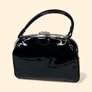 May include: A glossy black handbag with a curved handle and silver-toned hardware. The bag has a rectangular shape with rounded edges and a shiny, reflective surface. The handle is attached to the top of the bag with metal clasps.