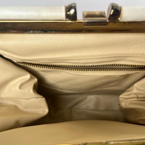 May include: The inside of a vintage handbag with a cream colored lining and a gold zipper. The handbag has a gold metal frame and a white top.