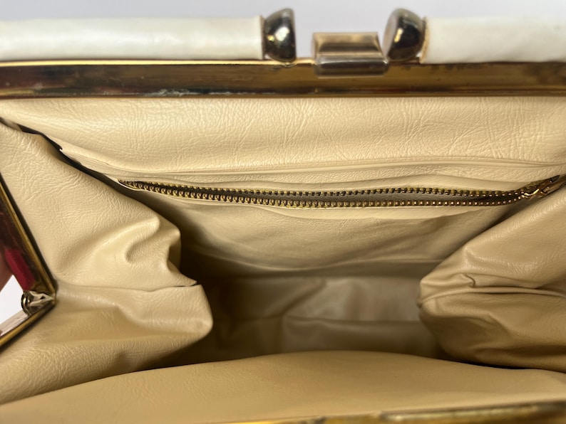 May include: The inside of a vintage handbag with a cream colored lining and a gold zipper. The handbag has a gold metal frame and a white top.