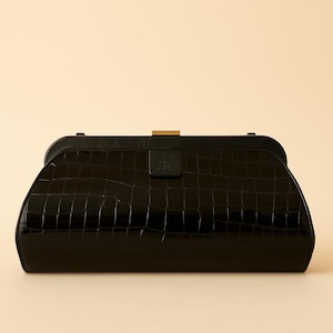 May include: A black clutch with a crocodile-textured pattern. The rectangular bag has a curved top and a gold-colored clasp. A small, embossed emblem is centered on the front.