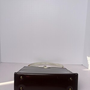 May include: A dark brown patent leather handbag with a white top handle and gold-toned hardware. The bag has a rectangular shape and a flat bottom.