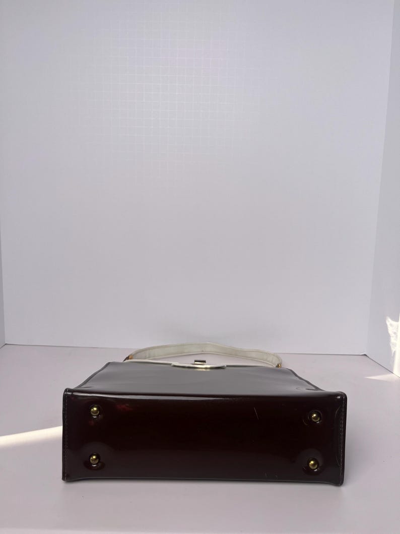 May include: A dark brown patent leather handbag with a white top handle and gold-toned hardware. The bag has a rectangular shape and a flat bottom.
