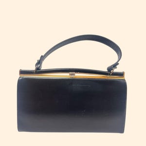 May include: A black rectangular handbag with a curved handle. The bag has a gold-colored frame and clasp. The handle is attached to the top of the bag with small metal attachments.