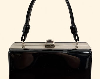 Puede incluir: Un bolso de mano rectangular de charol negro con acabado brillante. El bolso presenta un marco de metal plateado, una sola asa superior arqueada y un cierre de presión plateado en el centro del borde superior estructurado.