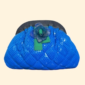 May include: A quilted blue clutch bag featuring a dark blue frame, a textured dot pattern, and a central decorative element consisting of a green ribbon strip topped with a translucent blue flower-shaped ornament.