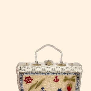May include: A white wicker handbag with a clear handle and gold-colored hardware. The front features nautical-themed embellishments, including a red ship's wheel, anchors, and a star. A decorative bow and strawberry are also present.