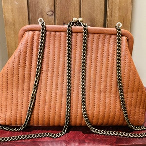 May include: A brown quilted clutch bag with a gold chain strap. The bag has a clasp closure and a slightly rounded shape.