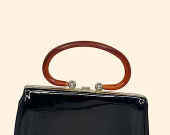 May include: A vintage black patent leather handbag with a rounded, tortoiseshell-colored handle. The rectangular purse has a silver clasp and a glossy finish, showcasing a classic retro design.