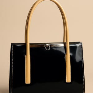 May include: A black patent leather handbag with tan handles. The rectangular bag has a silver clasp and stitching detail. The handles are arched and attached to the top of the bag. The bag is set against a neutral background.