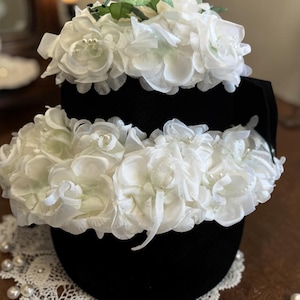 Puede incluir: Un sombrero negro adornado con capas de flores de tela blanca y pequeñas perlas. Hojas verdes se intercalan entre las flores. El sombrero se apoya en un tapete de encaje blanco, con perlas dispersas.