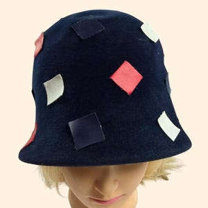 May include: A navy blue cloche hat adorned with colorful leather squares in red, white, and navy blue. The hat has a rounded crown and a slightly flared brim, offering a vintage aesthetic.