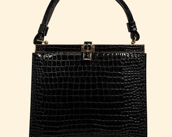 Puede incluir: Un bolso de mano negro estructurado con acabado brillante y patrón texturizado de piel de cocodrilo. El bolso cuenta con un asa superior, una silueta rectangular y un cierre de metal dorado centrado en el borde superior del marco.