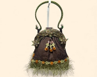 May include: A brown evening bag with a green braided handle and decorative fringe. The bag features floral embellishments, beaded accents, and small orange pom-poms. The bag is approximately 8 inches tall.