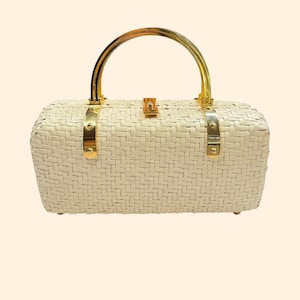 Puede incluir: Bolso de mano tejido de color crema con forma rectangular. Cuenta con un asa arqueada dorada y detalles metálicos dorados. El bolso tiene un cierre de broche y una textura tejida.