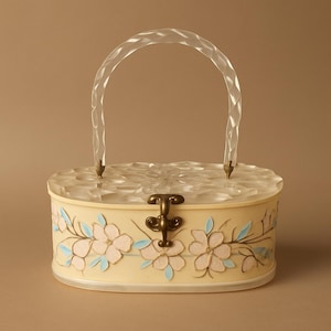 May include: A vintage-style, oval-shaped handbag with a clear, arched handle. The cream-colored body features floral designs in pink and blue. The clasp and handle attachments are brass-toned. The top of the bag has a textured, clear design.