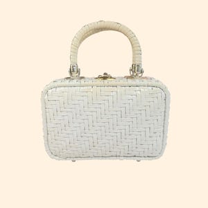 May include: A white woven handbag with a rounded handle. The rectangular bag features a textured, basket-weave design. The handle is a lighter shade of white and is attached to the bag with silver-coloured hardware. The bag has a small, gold-coloured clasp.