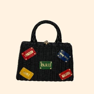 May include: Black woven handbag with a curved handle and a gold-colored clasp. The bag features colorful rectangular patches with the names of cities: London, Paris, Rome, Hong Kong, and Madrid. The patches have gold lettering.