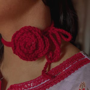 May include: A deep red crochet choker featuring a prominent, layered floral design at the center. The accessory is crafted from thick yarn and includes long, adjustable tie strings that hang down, allowing for a secure fit around the neck area.