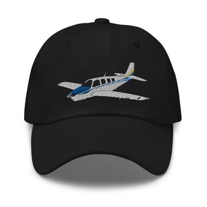 Könnte beinhalten: Eine schwarze Baseballkappe mit einem gestickten Bild eines kleinen, weißen Flugzeugs mit blauen und gelben Akzenten.