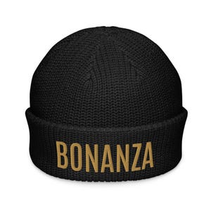 Könnte beinhalten: Schwarze Mütze mit dem Wort "BONANZA" in goldfarbenem Faden auf der Vorderseite gestickt. Die Mütze ist aus geripptem Strickmaterial gefertigt und hat einen gefalteten Saum. Eine warme und stilvolle Wintermütze.