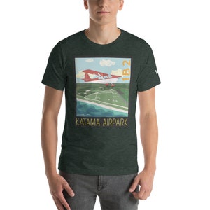 May include: A green t-shirt featuring a graphic of a red and white aircraft flying over a green field and blue water. The graphic also includes the text "Katama Airpark" and "1B2".