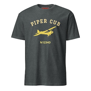 Puede incluir: Camiseta gris oscuro con un gráfico amarillo de un avión Piper Cub. El texto "PIPER CUB" está arqueado sobre el avión, con "N1234D" debajo. La camiseta está hecha de un material suave.
