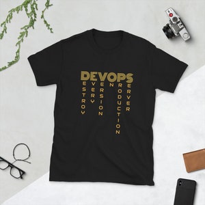 Puede incluir: Camiseta negra con un gráfico dorado que dice "DEVOPS" con las letras dispuestas para deletrear las palabras "VERSION", "SERVER", "TRY", "ROY", "PRODUCTION" y "ERROR".