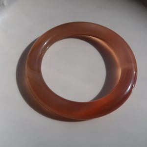 Può includere: Un braccialetto a cerchio traslucido color ambra. Il braccialetto circolare è realizzato in un materiale liscio e lucido e proietta un'ombra sulla superficie bianca su cui poggia. Il gioiello è un accessorio semplice ed elegante.