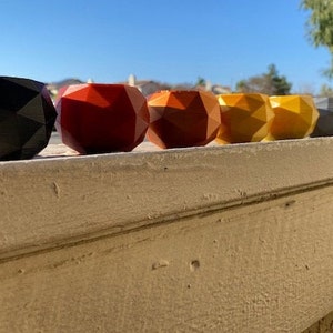 May include: A row of eight geometric planters in various colours, including black, red, orange, yellow, and grey. The planters are made of a smooth, textured material and are arranged in a line on a light brown surface.