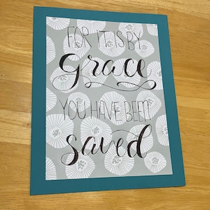 May include: A teal framed print with a gray background featuring a white floral pattern. The print has black calligraphy that reads "For it is by grace you have been saved."