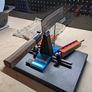 May include: A black knife sharpening system with a blue base and orange and red sharpening stones. The knife is being sharpened on the system.