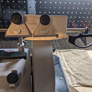 May include: A close-up of a knife sharpening system with a knife in the clamp. The system has two adjustable angle guides and a gray grinding wheel. The knife is a black handled pocket knife with a partially serrated blade.