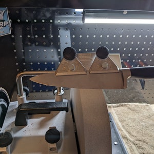 Peut inclure: Gros plan d'un système d'affûtage de couteau avec un couteau en place. Le système comporte deux guides métalliques avec des boutons noirs et un bras métallique avec une extrémité incurvée. Le couteau repose sur les guides et le bras maintient le couteau en place. Le système est fixé à une machine grise avec une meule abrasive brune.